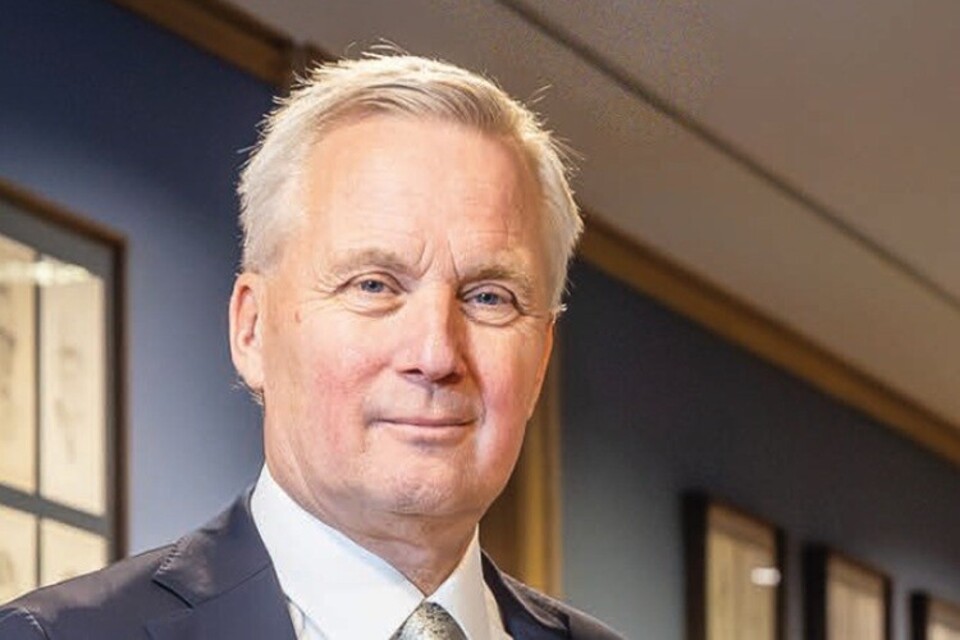 Man met grijs haar, blauw pak, wit overhemd en stropdas, glimlachend in een gang met ingelijste schilderijen.