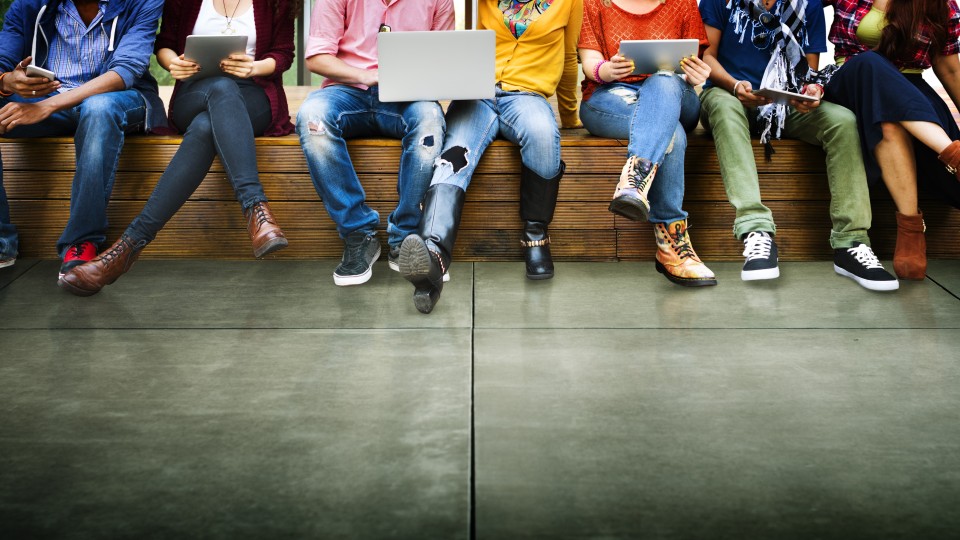 jongeren zitten op een rij. Alleen hun rom en benen zijn zichtbaar. In hun handen zie je laptops, tablets en mobiele telefoons