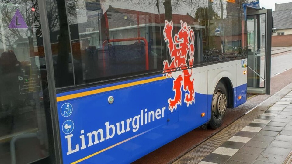 Blauwe en witte bus met het woord Limburgliner en een rood Limburgs wapen, geparkeerd bij een halte met open deur.