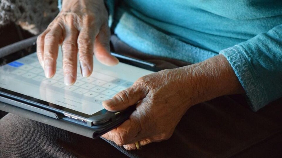 Handen van een oudere persoon die een tablet vasthouden en het scherm bedienen met een virtueel toetsenbord.