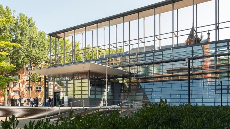 Moderne glazen gevel van het gerechtsgebouw met mensen bij de ingang en groen op de voorgrond.