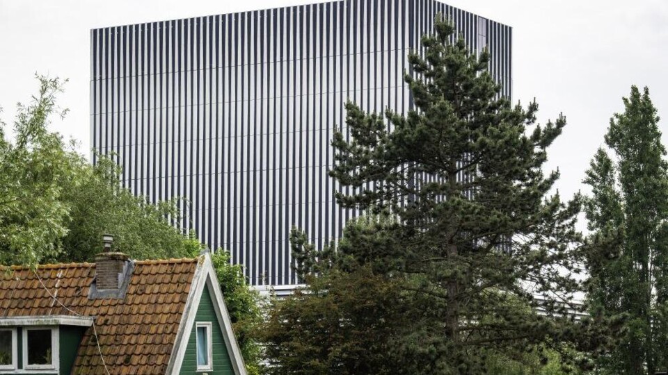Groot modern datacentergebouw met verticale strepen achter groene bomen en een huis met rood dak.