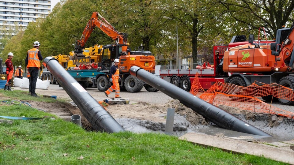 Bouwvakkers en graafmachines bij aanleg van warmtenet in woonwijk met grote zwarte buizen en bomen.