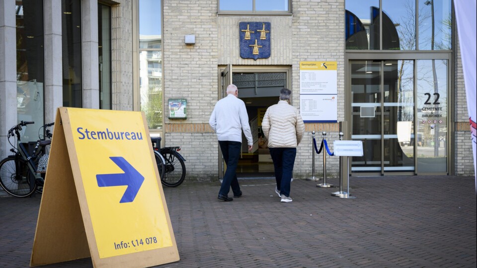 Twee mensen lopen een gebouw binnen met een bord 'Stembureau' en een blauw-geel bord bij de ingang.