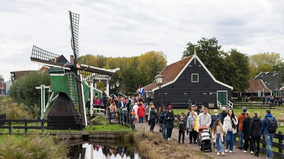 Drukke wandelende toeristen bij een groene windmolen en zwart houten huis aan water in Zaanse Schans.