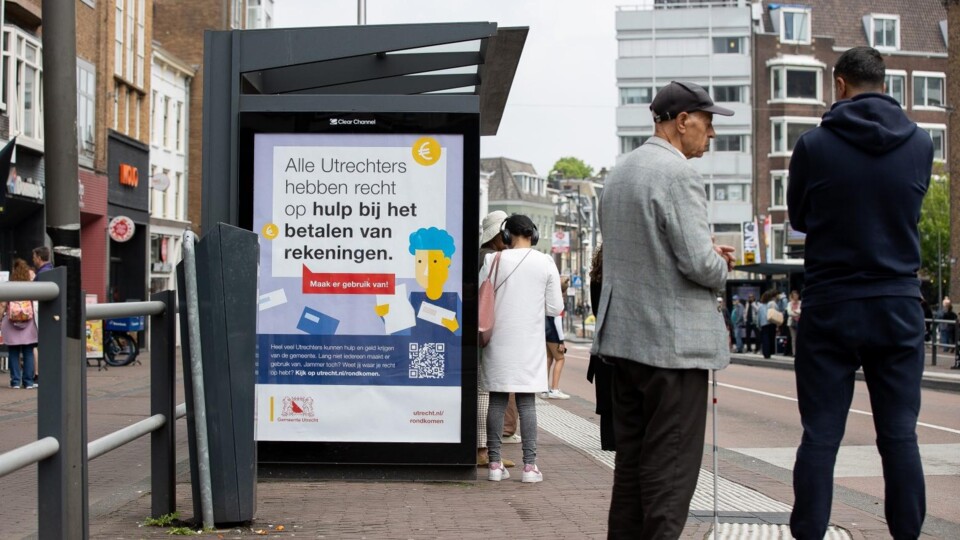 Poster bij bushalte in Utrecht met tekst over hulp bij het betalen van rekeningen en mensen op straat.