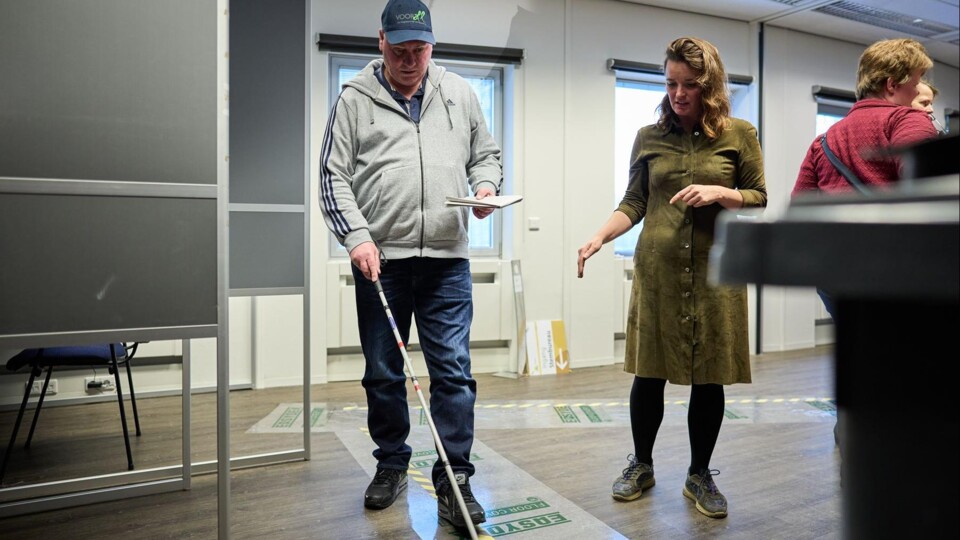 Man met witte stok en pet oefent lopen in een stemlokaal, vrouw wijst naar de grond, andere mensen op de achtergrond.