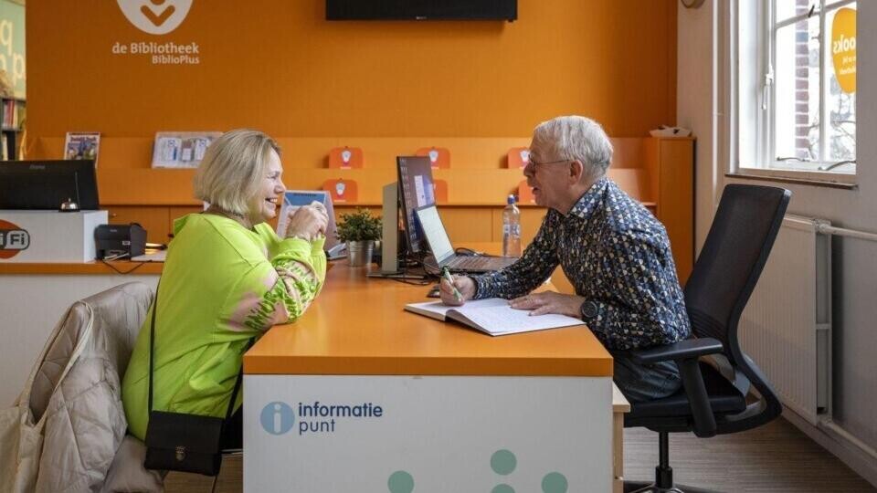 Vrouw en man praten aan tafel bij informatiepunt digitale overheid in bibliotheek met oranje muur.