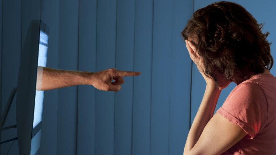 Hand wijst vanuit computerscherm naar vrouw die haar gezicht met handen bedekt, zittend aan bureau met toetsenbord.