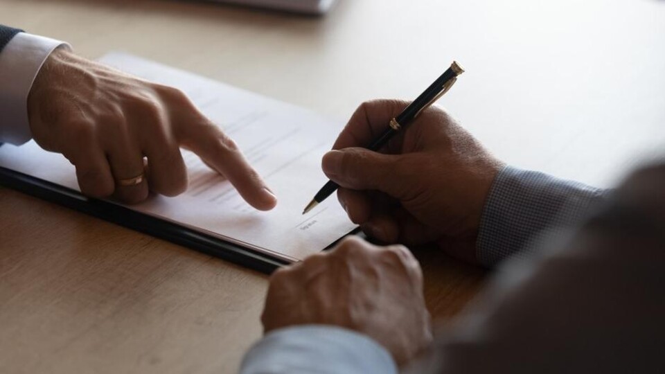 Handen van twee mannen, één wijst op contract, ander houdt pen om te tekenen op houten tafel.