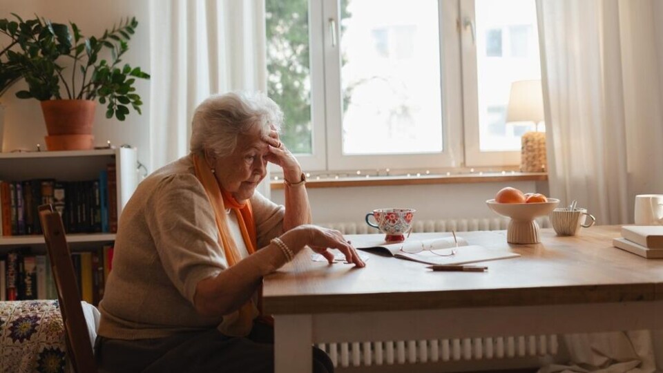 Oudere vrouw zit aan tafel met papieren, kopje thee en fruit, kijkt bezorgd en denkt na.