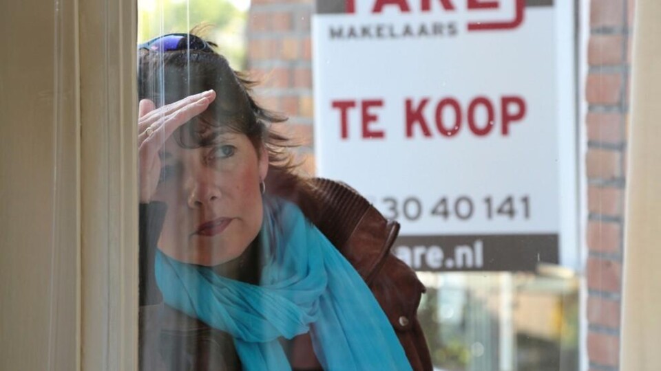 Vrouw met blauwe sjaal kijkt door raam naar 'Te Koop' bord van makelaarskantoor.