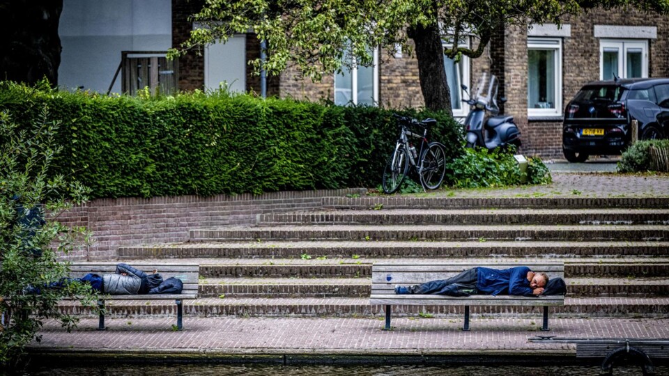 Twee mannen liggen te slapen op houten bankjes langs een gracht met trappen, fietsen en auto's op de achtergrond.