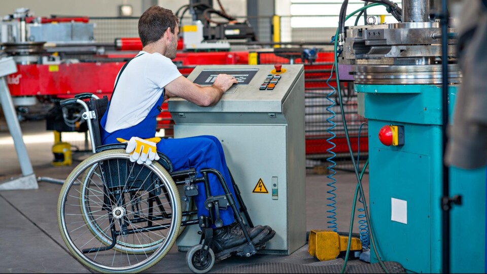 Man in rolstoel draagt blauwe werkbroek en bedient een bedieningspaneel van een machine in een fabriek.