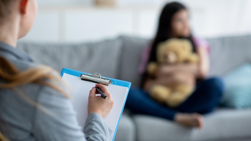 Jeugdzorgprofessional schrijft op clipboard terwijl kind met knuffel op bank zit, wazig op achtergrond.