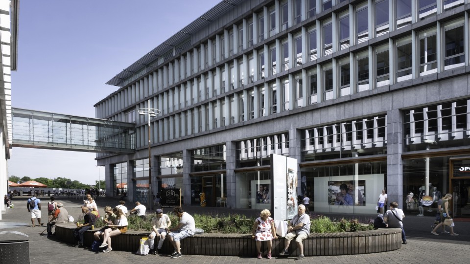 Mensen zitten in de zon op een muur voor het Maastrichtse stadskantoor in het winkel- en kantorencomplex Mosae Forum in het centrum van…