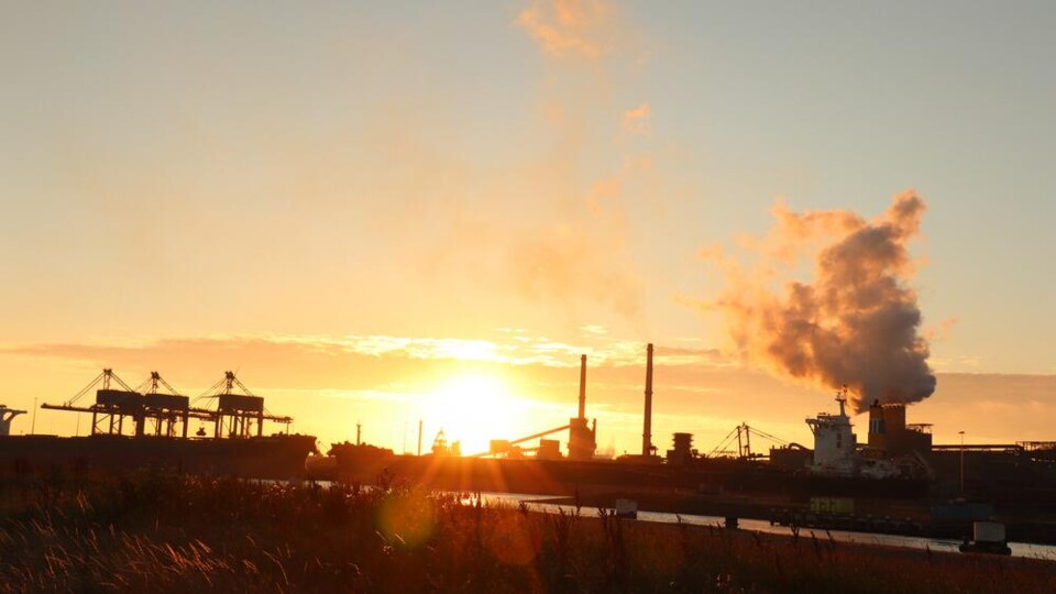 Zonsondergang bij Tata Steel fabriek met rookpluimen uit schoorstenen en silhouetten van industrie.