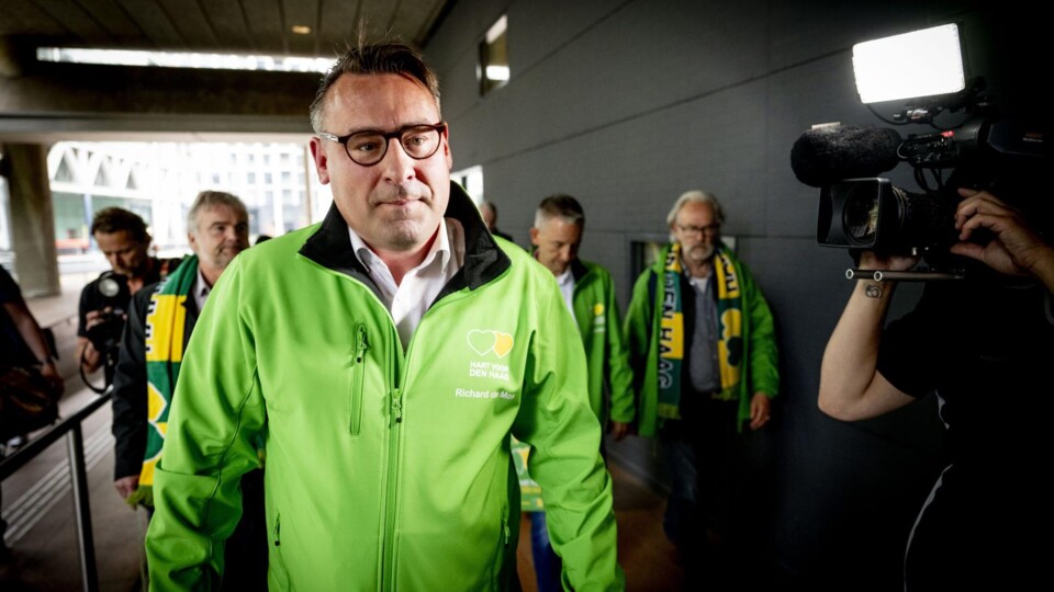 Man in groene jas met bril loopt voor een groep mensen, gefilmd door cameraman met licht.