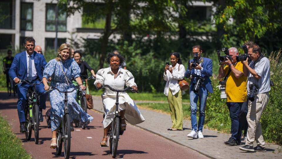 Koningin Máxima en wethouders fietsen op een fietspad in Amsterdam-Zuidoost, gefotografeerd door persfotografen aan de zijkant.