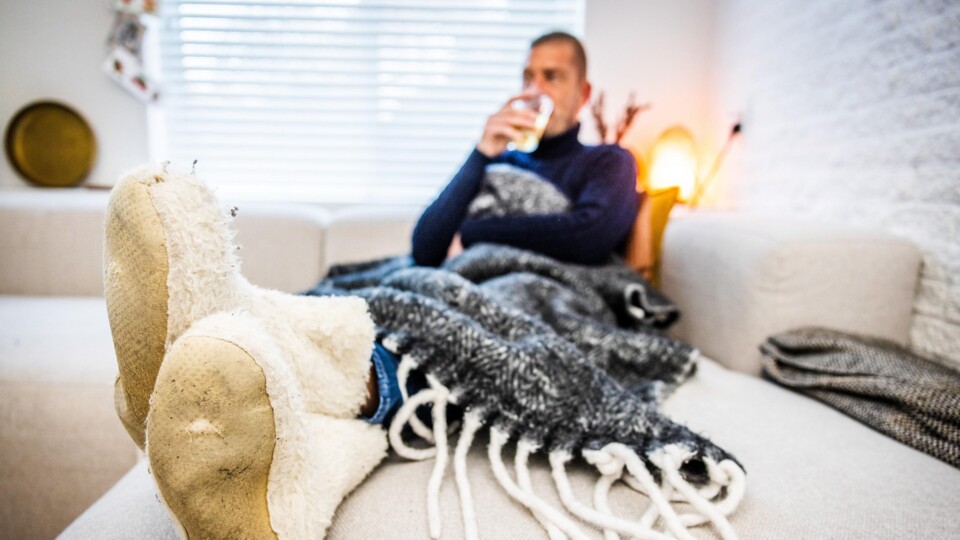 Man met warme sloffen en deken zit op bank en drinkt uit glas in woonkamer.