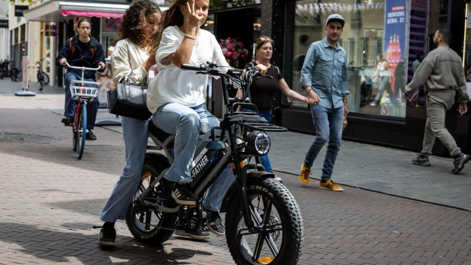 Twee vrouwen op een zwarte fatbike met dikke banden in een drukke winkelstraat met meerdere voetgangers
