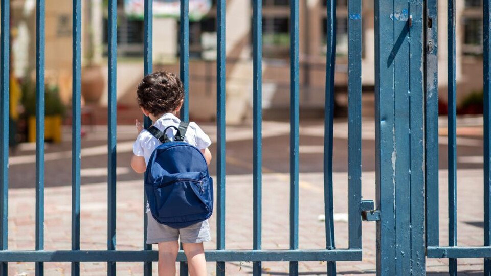 Jong kind met blauwe rugzak staat voor een gesloten blauwe metalen schoolpoort, gezien van achteren.