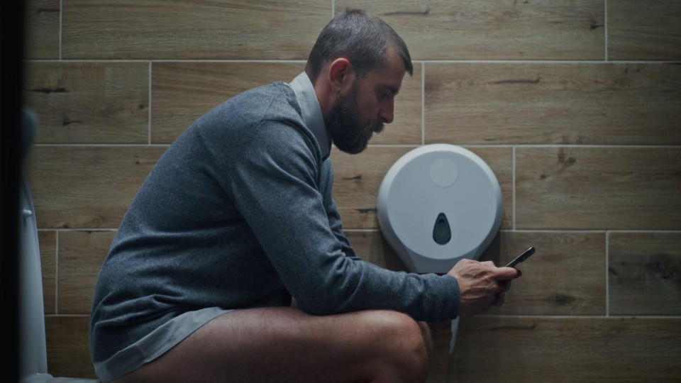 Man met broek op de enkels zit op de wc op zijn smartphone
