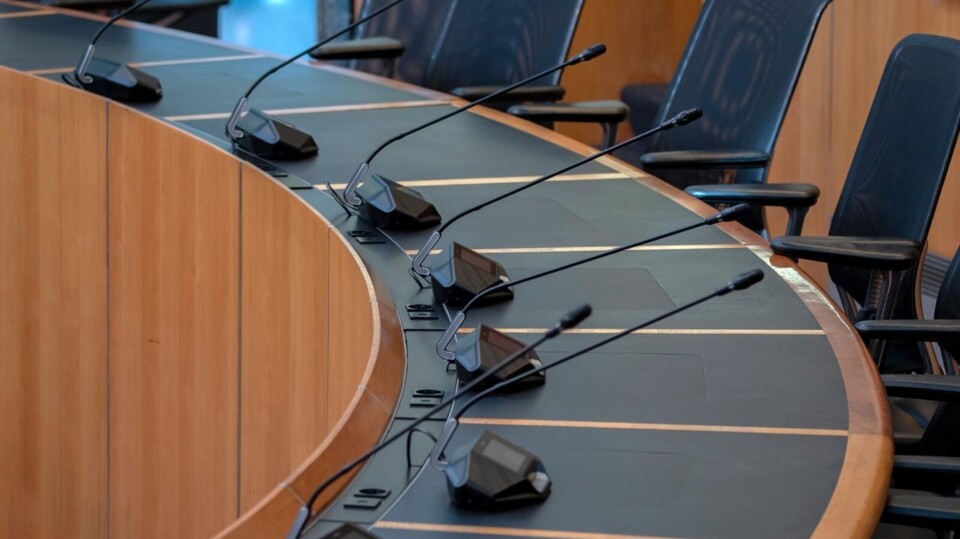 Ronde tafel met microfoons en zwarte stoelen in een lege gemeenteraadsvergaderruimte.