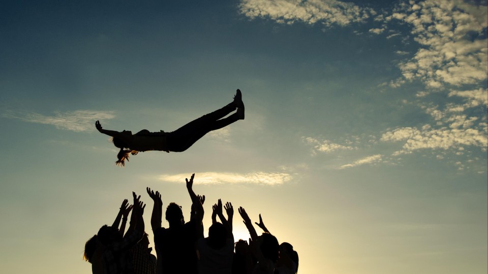 Een groep mensen vangt gezamenlijk een vrouw op die uit de lucht komt vallen