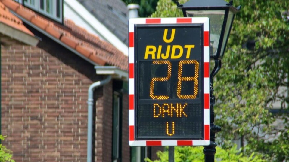 Snelheidsdisplay met tekst 'U RIJDT 28 DANK U' langs een straat met huizen en bomen.