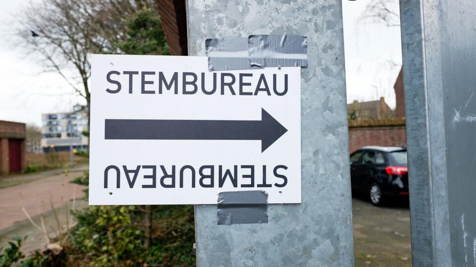 Wit bord met zwarte tekst 'STEMBUREAU' en een pijl naar rechts, bevestigd met ducttape aan een metalen paal buiten.