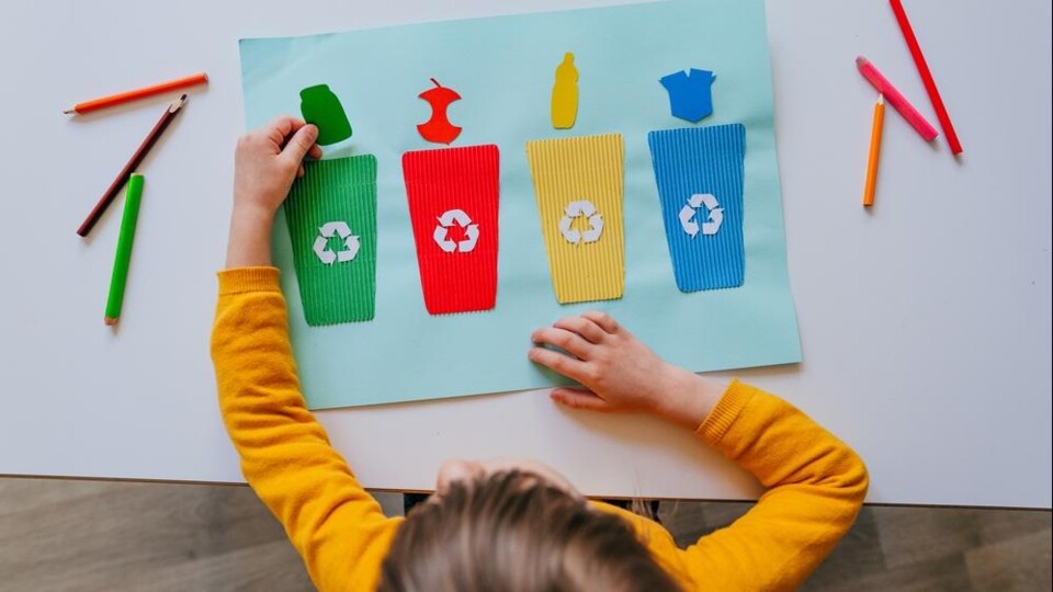 Kind met gele trui plakt papieren recyclebakken in groen, rood, geel en blauw met afvalsymbolen op lichtblauw papier.