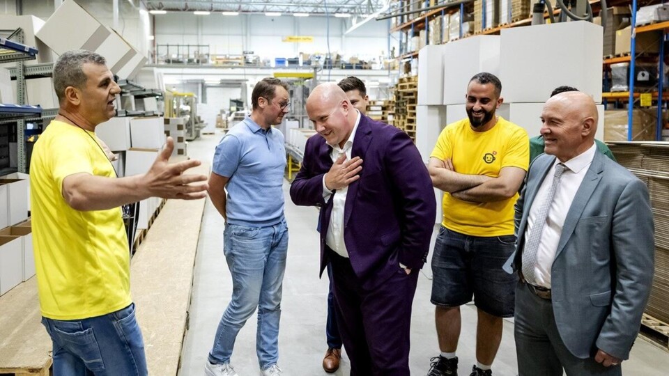 Man in paars jasje wordt rondgeleid door werknemers in gele shirts in industriële werkplaats.