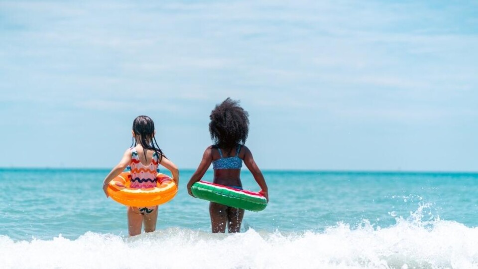 Twee kinderen met zwembanden lopen het water in bij een strand op een zonnige dag, gezien van achteren.