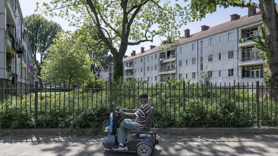 Man op scootmobiel rijdt langs sociale huurwoningen met bomen en hekken in de buurt.