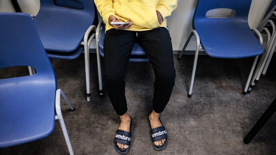 Man in slippers, hoofd niet te zien, zit op opgestapelde stoelen, kijkt op telefoon