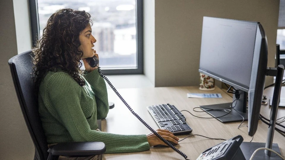 Vrouw in groen shirt werkt aan callcenter bureau met koptelefoon, telefoon en computermonitor.