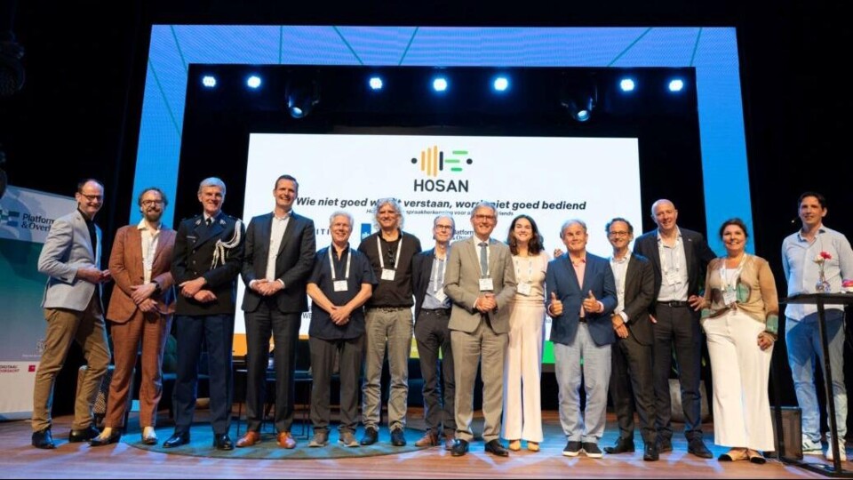 Groepsfoto van deelnemers aan het HOSAN congres op het podium met een groot scherm op de achtergrond met het HOSAN logo en tekst.