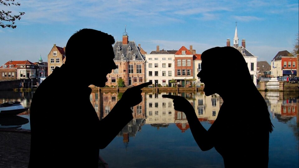 Silhouetten van een man en een vrouw die naar elkaar wijzen met oude gebouwen en waterreflecties Maassluis op de achtergrond.