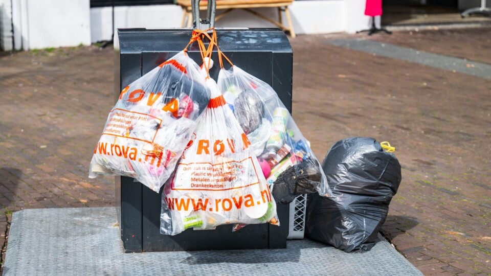 Drie doorzichtige afvalzakken met oranje ROVA-logo hangen aan een zwarte afvalcontainer op straat.
