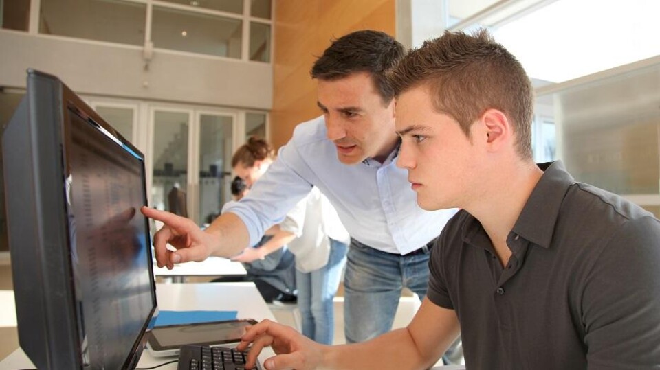 Begeleider helpt jongeman bij computerwerk in kantooromgeving met meerdere personen op achtergrond