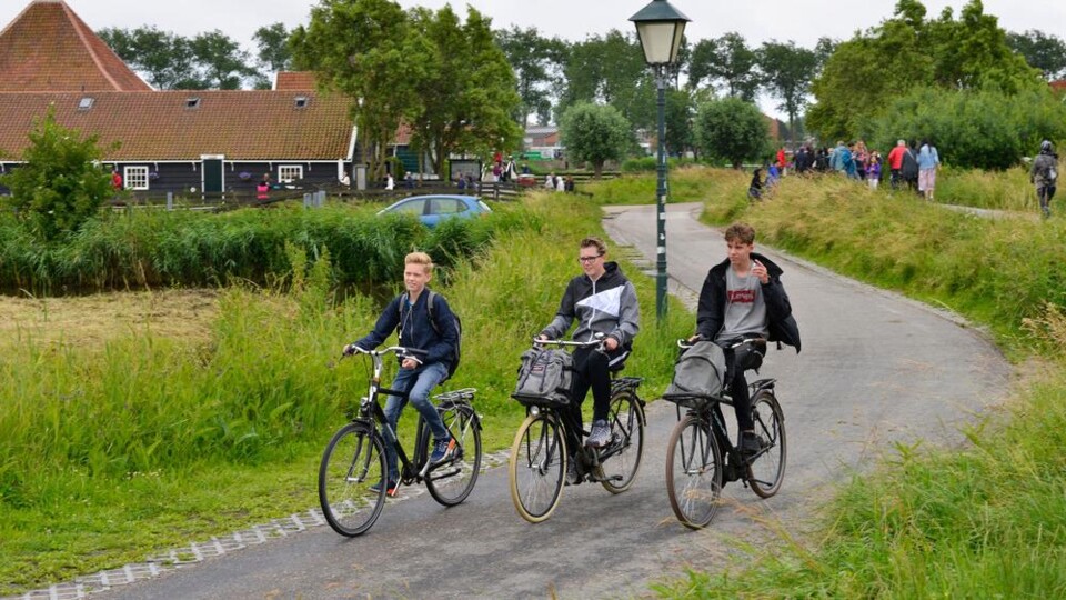 Drie jongens fietsen op een smal landweggetje met groene begroeiing en huizen op de achtergrond.
