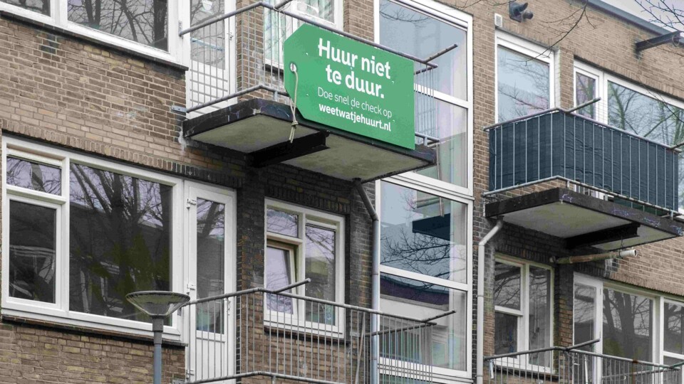 Groen bord met tekst 'Huur niet te duur' aan balkon van een appartementencomplex met bakstenen gevel.