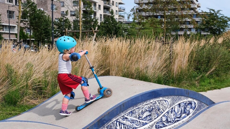 Jong kind met blauwe helm en kniebeschermers op blauwe step in speeltuin met gras en gebouwen op achtergrond