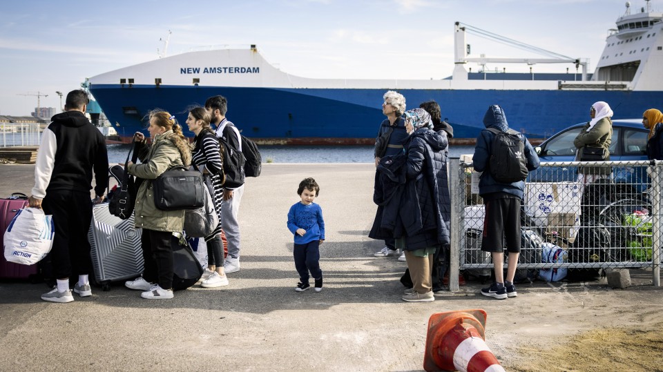 Een kleuter staat tussen 2 groepen asielzoekers, op de achtergrond staat een cruiseschip.