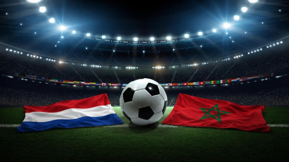 Voetbal op grasveld tussen Nederlandse en Marokkaanse vlaggen in een verlicht stadion met publiek.