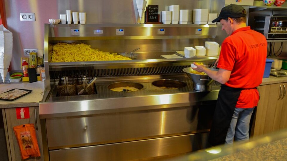 Medewerker in rood shirt en zwarte pet bereidt friet bij een frituur met grote hoeveelheid friet en friteuse in een snackbar.