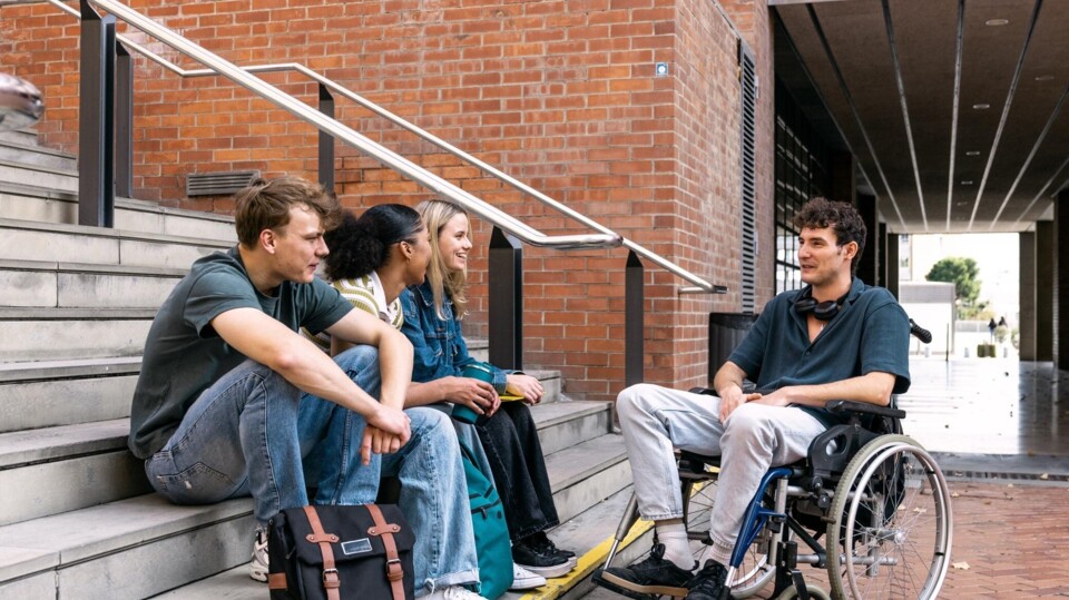 Groep studenten zit op trappen buiten gebouw, één student in rolstoel, allen in gesprek met elkaar.