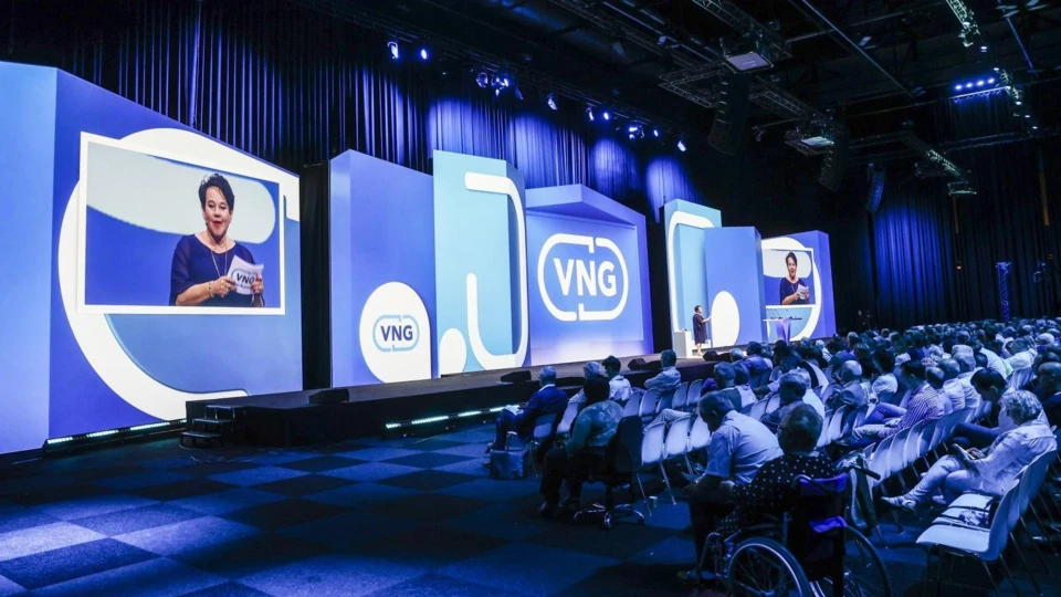 Spreekster presenteert op VNG Algemene Ledenvergadering met VNG-branding en deelnemers in zaal