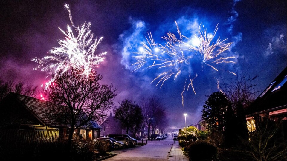Blauwe en witte vuurwerkexplosies boven een donkere woonstraat met geparkeerde auto's en bomen.